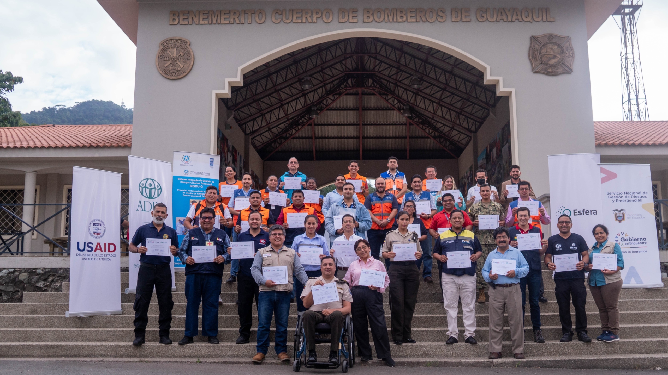 A group of 37 people holding certificates stand on the steps outside a building. One person is in a wheelchair. Rollup banners for USAID, ADRA and Sphere are visible. The letters on the building read “Benemerito Cuerpo de Bomberos de Guayaquil”