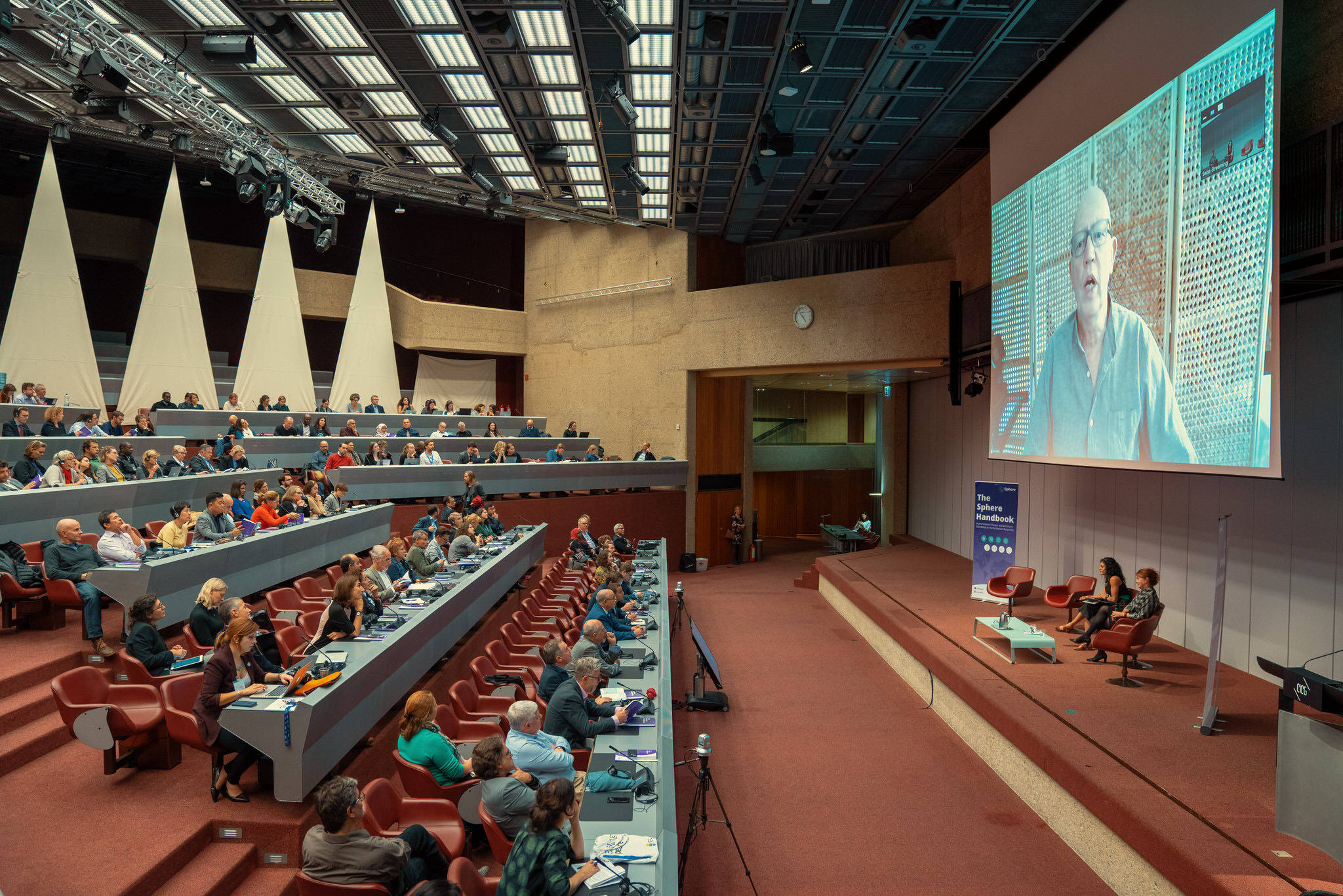 Participantes del lanzamiento del Manual Esfera en Ginebra escuchan a Peter Walker dar el discurso de apertura en una transmisión de vídeo en directo