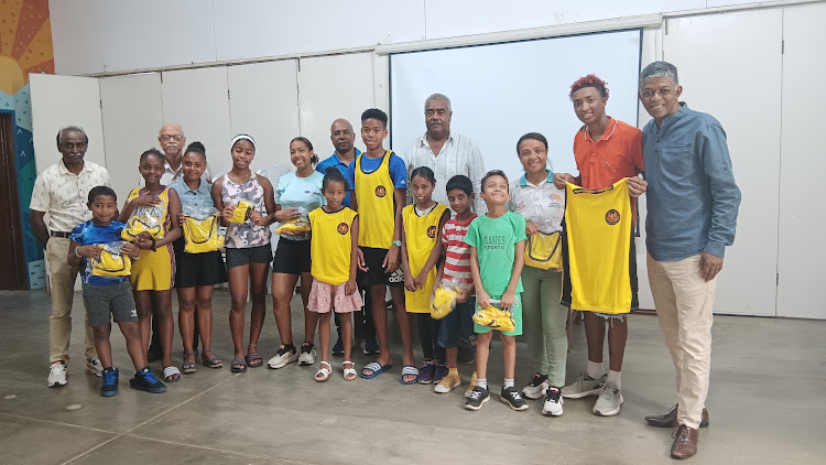 A group of adults and children stand indoors posing for a photo, some holding yellow sports jerseys and snack packs.